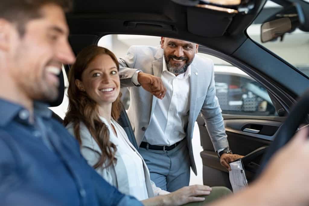 African salesman showing new car to young couple in showroom. Happy man and woman choosing auto in car dealership while cardealer explaining the new technology.