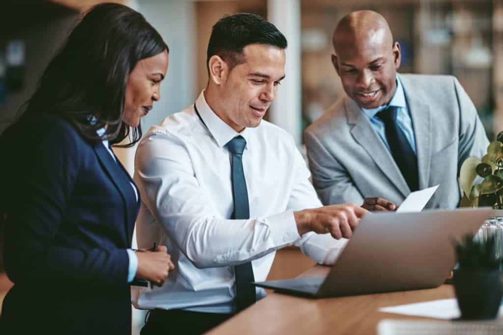 Diverse group of smiling businesspeople reading paperwork together while working on a laptop at a table in an office