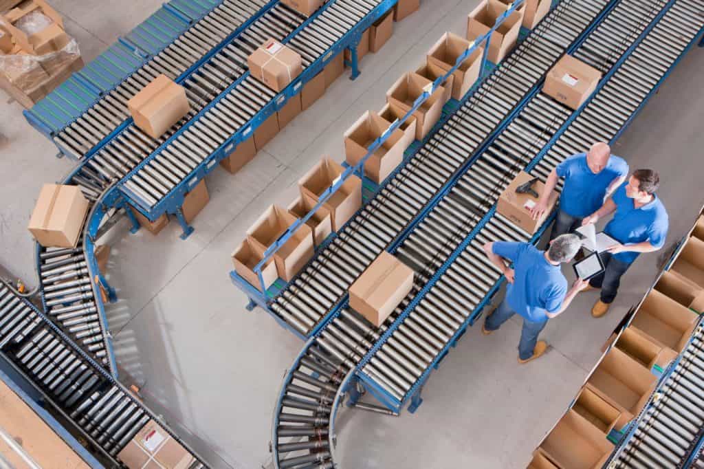 Top view of workers with papers and a digital tablet having a discussion among boxes laid on conveyor belts at a distribution warehouse.