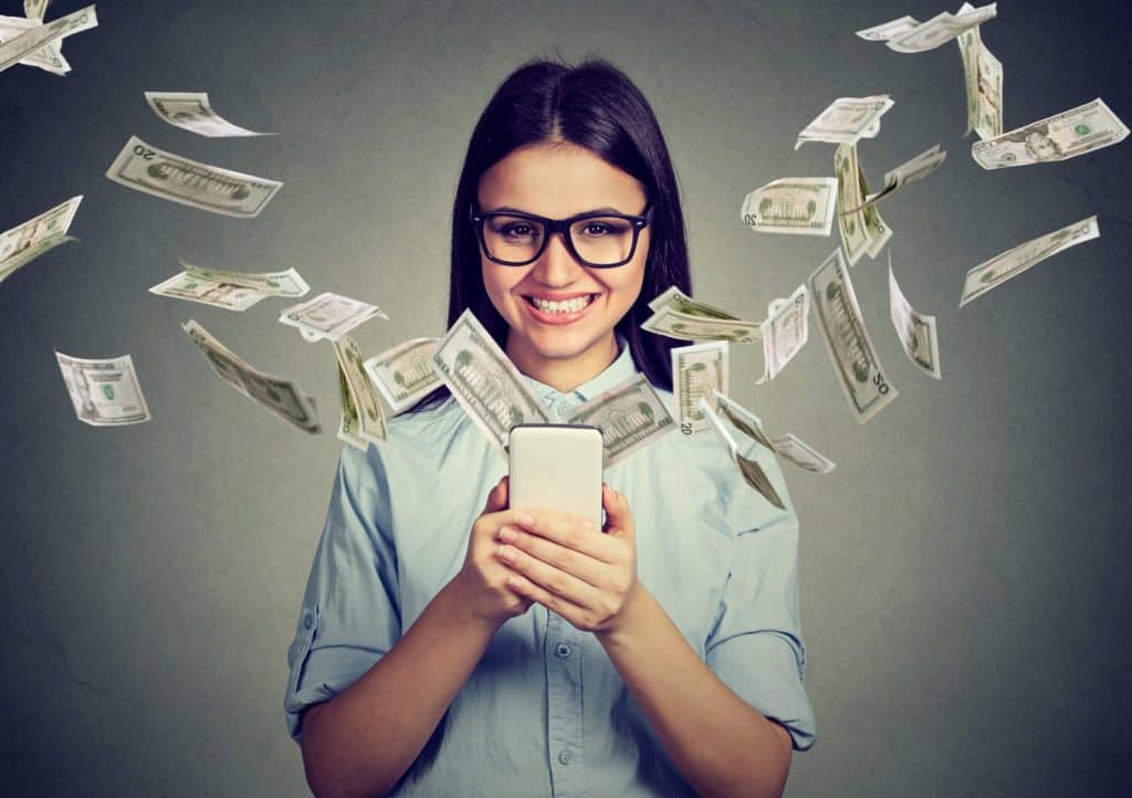 Online banking money transfer, e-commerce concept. Happy young woman in glasses using smartphone with dollar bills flying away from screen isolated on gray wall background.